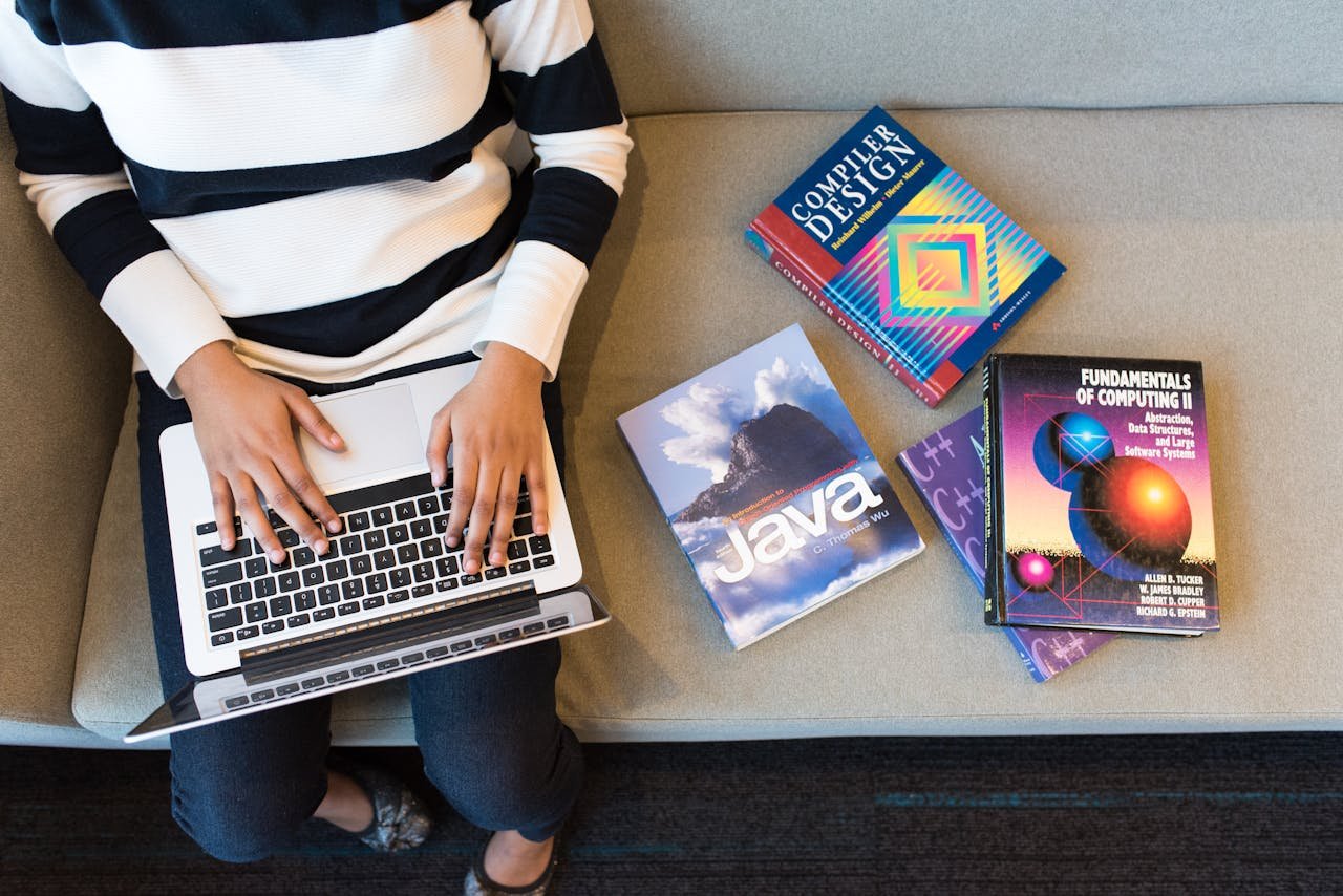 portfolio-05 Woman using laptop on sofa, surrounded by programming books, learning coding.